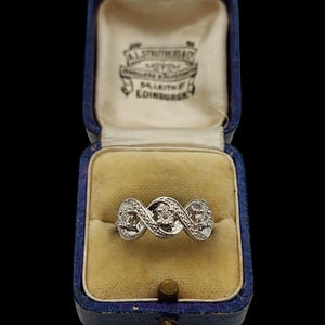 Könnte beinhalten: Ein silberfarbener Ring mit drei kreisförmigen Fassungen und kleinen eingelassenen Steinen, präsentiert in einer rechteckigen blauen Samtschmuckschatulle mit cremefarbenem Innenfutter, das den Text A.L. STRUTHERS & CO. JEWELLERS & SILVERSMITHS 54 LEITH ST EDINBURGH zeigt.