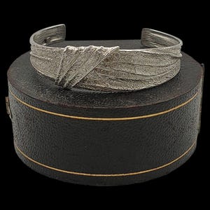 May include: A silver cuff bracelet with a textured, leaf-like pattern. The bracelet is displayed on a black, round box with gold edging, against a black backdrop. The cuff has an open design. The bracelet is likely made of silver.