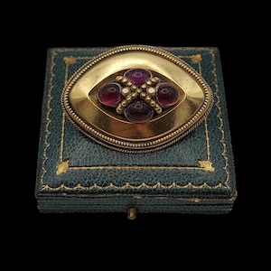 May include: An oval gold brooch featuring four deep red cabochon stones arranged in a cross pattern, accented by small gold beads. The brooch rests on a square, dark teal textured box with gold decorative trim along the edges and corners.