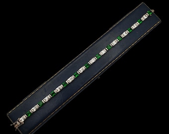 May include: A silver bracelet featuring alternating emerald green and clear rectangular stones. The bracelet is presented on a dark blue jewellery box against a black backdrop. The clasp is visible.