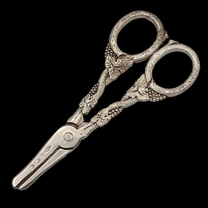 Peut inclure: Gros plan sur une paire de ciseaux anciens en argent, avec des détails ornés de vignes et de feuilles. Les ciseaux ont des trous ovales pour les doigts et un design articulé. Fond noir.