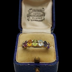 May include: A gold ring featuring seven oval-cut gemstones in a row, including purple, orange, lime green, light blue, and dark blue stones. The ring is displayed inside a blue velvet-lined jewellery box with the text A.L. STRUTHERS JEWELLERS EDINBURGH.