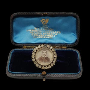 Op de afbeelding: Een antieke broche met een portret van een man, omringd door heldere edelstenen. Gepresenteerd in een zwarte leren etui met een blauwe fluwelen binnenkant. De etui draagt de tekst "W.L. LAWSON GOLDSMITHS & JEWELLER" en het adres.