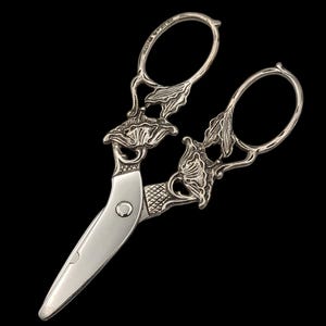May include: Silver-toned scissors with ornate floral detailing. The handles and the area where the blades meet feature intricate leaf and flower designs. The blades are smooth and shiny, set against a black background.