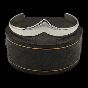 May include: A silver cuff bracelet with a distinctive chevron shape. The bracelet sits on a dark brown box with gold trim. The background is black. The bracelet's design is a modern take on a classic style.