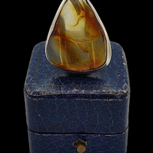 May include: A silver ring featuring a large, amber-colored stone with internal patterns. The ring is set on a blue jewelry box with a gold-colored clasp, against a black background. The amber stone has a unique, organic shape.