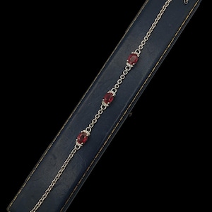 Può includere: Un bracciale a catena color argento con tre pietre sfaccettate rosso scuro di forma ovale. Ogni pietra è incastonata in una piccola montatura decorativa color argento, e il bracciale include una chiusura a moschettone e una breve catenella.