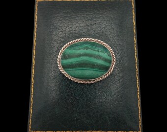 May include: Oval malachite brooch with a silver rope-like border. The green stone features natural banding. The brooch is set on a dark green leather-like surface with gold trim, against a black background.