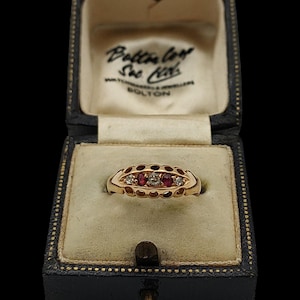 May include: A gold ring with three gemstones, two red and one clear, set in an openwork design. The ring is displayed in a vintage black jewelry box with the text "Bolton" visible.