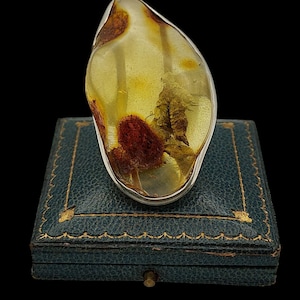 May include: A large, amber-colored ring with a silver band. The amber has inclusions of red and brown, set on a vintage teal jewelry box with gold trim. The ring's shape is organic and irregular.
