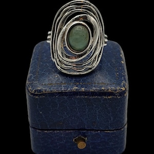Puede incluir: Anillo de plata con una piedra preciosa verde ovalada en el centro. El anillo tiene un diseño único, con un patrón en espiral en capas alrededor de la piedra. Se muestra sobre un joyero azul sobre un fondo negro.