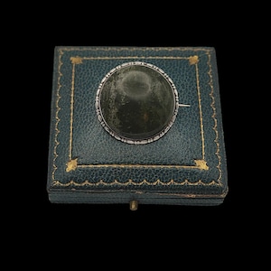 May include: A round, dark green brooch with a silver-colored filigree border, resting on a teal jewelry box with gold trim. The brooch has a pin on the back. The box is closed and has a small gold-colored clasp.