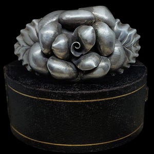 May include: A silver rose brooch with a detailed, textured design. The rose is made up of many small, rounded petals.
