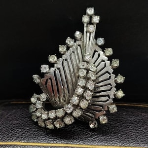 Puede incluir: Un broche de plata con un diseño geométrico y brillantes de cristal transparente. El broche tiene forma de hoja o flor estilizada.