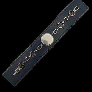 May include: A silver-toned watch with a round face and a delicate chain featuring floral and teardrop-shaped links. The watch is displayed on a blue leatherette box with a black background.