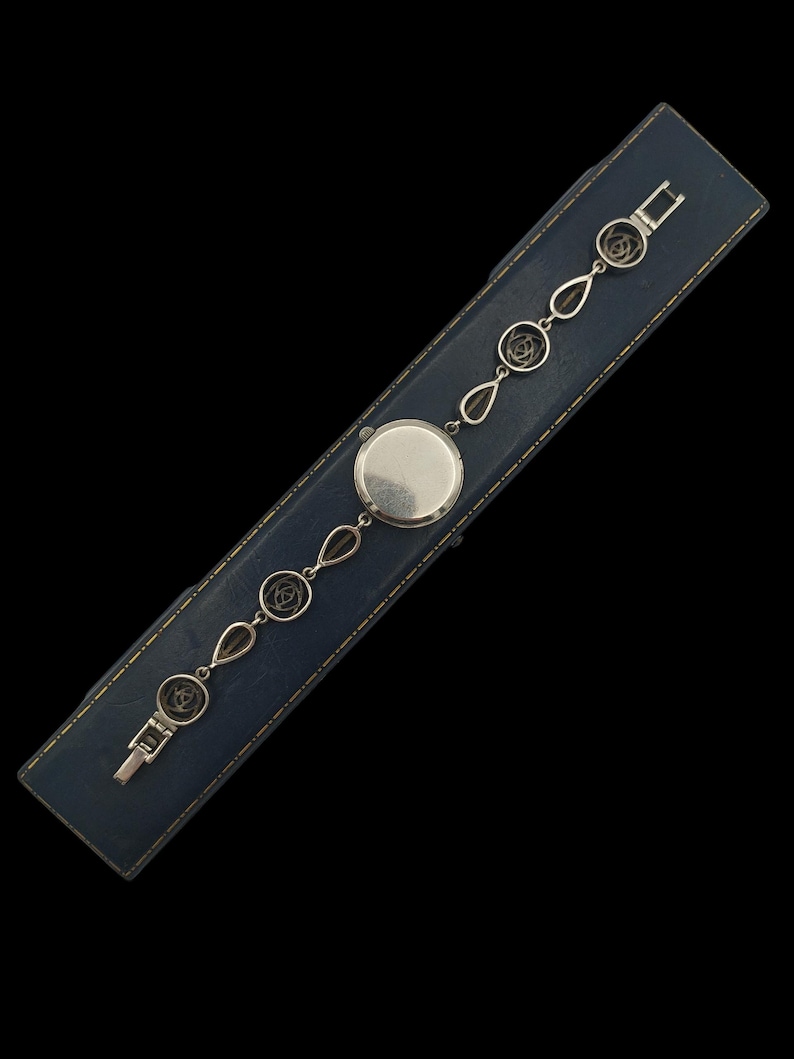 May include: A silver-toned watch with a round face and a delicate chain featuring floral and teardrop-shaped links. The watch is displayed on a blue leatherette box with a black background.