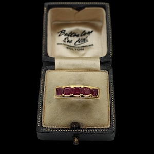 May include: A gold ring with five rectangular, deep red gemstones set in a row. The ring is displayed in an open, vintage black jewelry box with a cream-colored interior and the text "Bolton" on the inside lid.