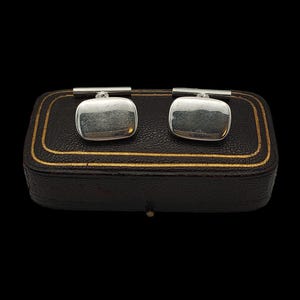 May include: Two silver cufflinks with a square, softly curved face, presented in a dark brown leather box with gold edging. The cufflinks have a textured finish and a silver bar. The box is approximately 10 cm long.