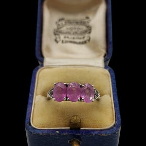 May include: A silver ring featuring three oval-cut pink gemstones set in a row, displayed inside a blue velvet-lined jewelry box with the text J. STRUTHERS, WATCHMAKER, EDINBURGH printed on the inner lid lining in black ink.