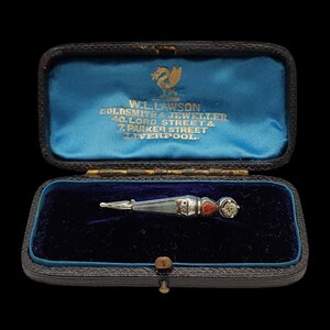 May include: An antique silver brooch with a geometric design, set with a red stone and a clear, faceted stone. The brooch is displayed in a black leather box with a blue velvet interior. The box has gold lettering that reads "W.L. LAWSON GOLDSMITH & JEWELLER".