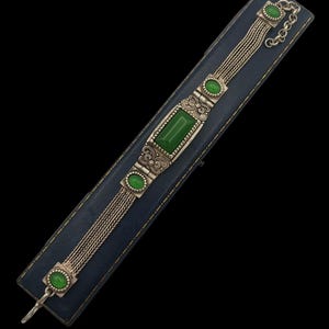 May include: A silver bracelet with a chain link design and green gemstone accents. The bracelet is displayed on a blue velvet box.