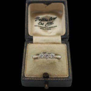 May include: A silver ring with three clear gemstones displayed in an open, vintage black jewelry box. The box has gold trim and a cream-colored interior. The text "Bolton Ltd" is visible on the box's interior.