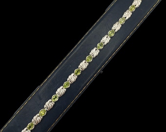 May include: A silver bracelet featuring oval, light green gemstones and decorative silver links. The bracelet is presented on a dark blue jewellery box against a black backdrop. The gemstones are evenly spaced along the bracelet.