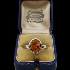 Puede incluir: Un anillo de plata con una piedra de ámbar translúcida de forma ovalada engastada en un bisel liso. El anillo se muestra dentro de un joyero azul oscuro con bisagras, forro de terciopelo color crema y texto impreso en la tapa interior.