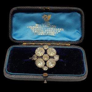 May include: An antique diamond brooch in a velvet-lined box. The brooch features a cluster of round-cut diamonds. The box is blue with gold lettering that reads "W.L. Lawson Goldsmith & Jeweller 40. Lord Street & 7. Parker Street Liverpool."