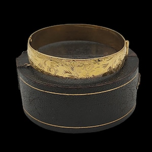 May include: A gold-coloured bangle bracelet with an engraved floral pattern. The bracelet is resting on a black, round jewellery box with a gold trim. The background is black.