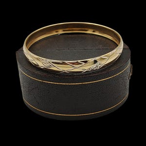 Op de afbeelding: Een gouden armband met een geometrisch ontwerp. De armband ligt op een donkerbruine, ronde doos met een gouden rand. De achtergrond is zwart.