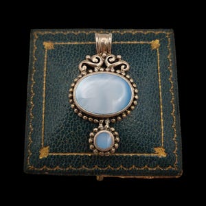 May include: A silver pendant featuring a large oval light blue cabochon stone and a smaller circular light blue cabochon stone below it. The silver setting includes decorative scrollwork at the top and a beaded border surrounding both stones.