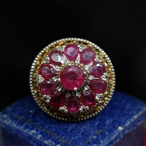 May include: A silver ring featuring a large, round, red gemstone encircled by smaller, white gemstones. The ring boasts a detailed, ornate design.