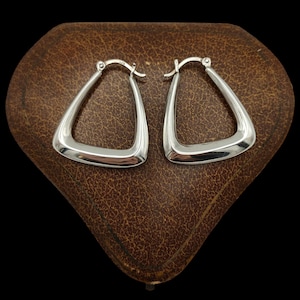 May include: A pair of polished silver hoop earrings with a distinct triangular shape. Each earring features a smooth, reflective surface and a hinged clasp mechanism at the top, showcasing a clean, metallic finish throughout the entire design.