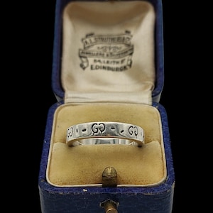 Op de afbeelding: Een zilveren ring met een herhalend logopatroon, gepresenteerd in een vintage blauwe sieradendoos met een gele satijnen binnenkant. De doos is open en toont het ontwerp van de ring.