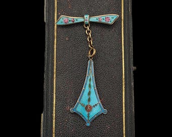 May include: A vintage brooch with a bow-shaped top and a pendant. The brooch features a light blue enamel design with pink floral accents and a gold chain. The pendant is a light blue enamel with a red center and green accents, set on a black leather box.
