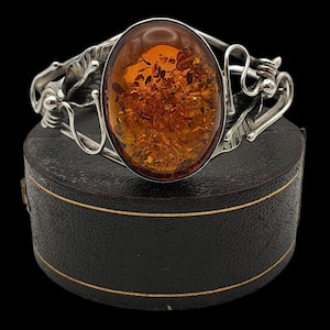 May include: A silver cuff bracelet with an oval amber stone. The amber is a warm, translucent orange color with inclusions. The silver setting has an ornate, flowing design. The bracelet is displayed on a black box.