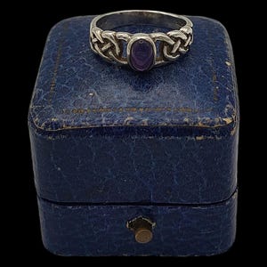 May include: A silver ring featuring a Celtic knot design, with an oval purple gemstone at its centre. The ring rests on a vintage blue jewellery box with a brass clasp, set against a black backdrop.