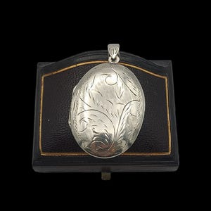 Puede incluir: Colgante ovalado de plata con un diseño floral grabado. El colgante se muestra en una caja negra con ribete dorado. Tiene un pequeño lazo para una cadena. El fondo es negro.