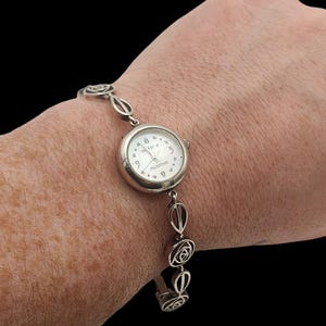 May include: Silver-toned wristwatch with a white face and delicate floral-inspired links. The watch face displays the brand name "The Itts". The band features oval and rose-shaped links, creating an elegant and feminine design.