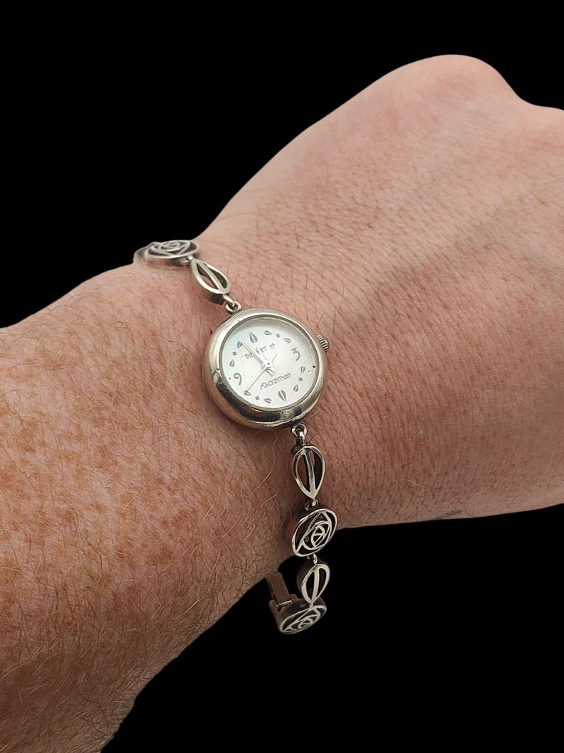 May include: Silver-toned wristwatch with a white face and delicate floral-inspired links. The watch face displays the brand name "The Itts". The band features oval and rose-shaped links, creating an elegant and feminine design.