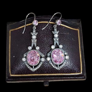Op de afbeelding: Een paar zilveren oorhangers met roze ovale stenen en heldere strass steentjes. De oorhangers hangen aan een bruine fluwelen doos.