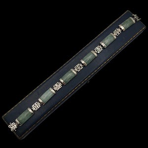 May include: A jade bracelet with cylindrical jade stones and silver-toned connectors. The bracelet is displayed on a blue, rectangular box against a black background. The connectors feature an openwork design.