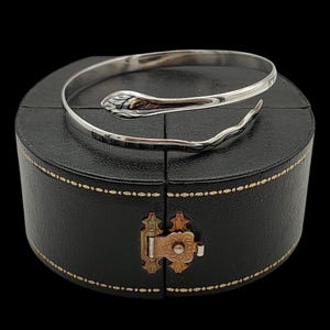 Puede incluir: Un brazalete de metal plateado con forma de serpiente enroscada. La pulsera presenta una cabeza lisa y pulida y un cuerpo texturizado y segmentado que se envuelve en un diseño en espiral. El metal tiene un acabado reflectante en toda la pieza.
