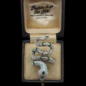 May include: A silver snake ring with green and blue enamel detailing, presented in a vintage jewellery box. The snake's head is light blue, and the body is adorned with gemstones. The box is black with gold lettering.