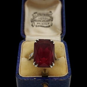 Puede incluir: Un anillo de plata con una gran piedra roja oscura rectangular facetada sujeta por cuatro garras. El anillo se presenta dentro de un joyero azul con bisagras, forro interior de color crema y una etiqueta impresa que dice A.L. STRUTHERS EDINBURGH.