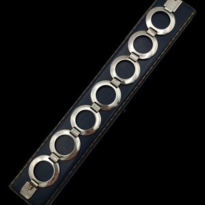 Könnte beinhalten: Ein silberfarbenes Gliederarmband bestehend aus einer Reihe von sechs kreisförmigen Ringen, die durch kleine, texturierte rechteckige Segmente verbunden sind. Das Armband liegt auf einem dunkelblauen, rechteckigen Etui mit Goldrand.