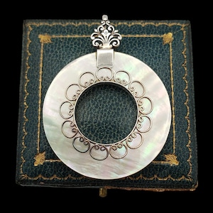 May include: A circular pendant featuring a flat, iridescent white mother-of-pearl disc. The centre of the disc is adorned with a silver filigree overlay consisting of interconnected scrolls. A silver decorative bail sits at the top of the pendant.