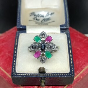 Peut inclure: Une bague en argent avec une pierre noire au centre et des pierres vertes et roses autour. La bague est sertie dans un motif floral avec des pierres noires autour des bords.