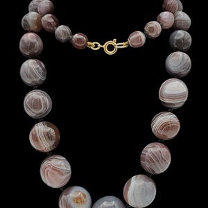 May include: A necklace composed of round, polished agate beads featuring concentric bands of brown, grey, and white. The beads are strung on a dark cord and secured with a circular gold-toned metal spring ring clasp at the top centre.